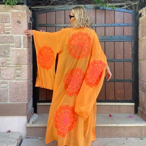 May include: An orange kimono-style robe with large, circular, floral-like patterns in a brighter orange hue. The robe has wide sleeves and a flowing design. The person is wearing sunglasses.