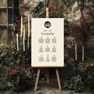 May include: A vertical rectangular wedding seating plan sign on a wooden easel. The cream-coloured sign features a dark blue circular logo with two swans, the text Sarah & Jack, The Seating Plan, and nine numbered tables with guest names.