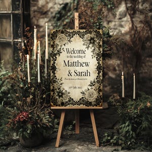 May include: A wooden easel displays a vintage-style wedding welcome sign. The sign features the text "Welcome to the wedding of Matthew & Sarah" with floral and lace-like border details. The date "22nd July 2027" is also visible.