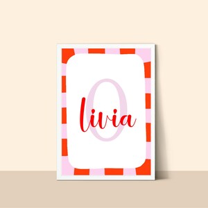 May include: A framed print featuring the name "Livia" in red script. The name is centered on a white background with a large, pale pink letter "O" behind it. The frame has a pink and orange striped border.