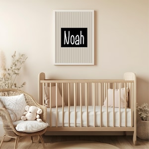 May include: A framed print with the name "Noah" in white letters on a black background, set against a beige and white striped pattern. The print hangs above a wooden crib in a nursery setting. A wicker chair with a teddy bear and pillows is in the foreground.