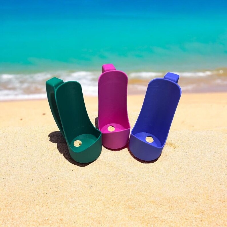 May include: Three colorful plastic cup holders in green, pink, and blue. The holders are designed to fit around a standard cup and have a hole in the bottom for drainage. The holders are sitting on a sandy beach.