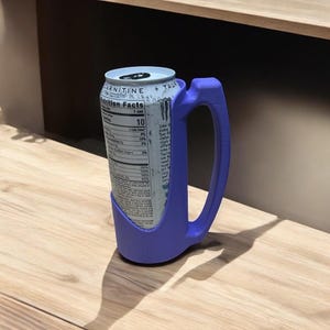 May include: A purple 3D printed can holder with a handle. The holder is designed to fit a standard 12-ounce can. The can is a white and blue energy drink can with the words "Carnitine + Taurine" and "Nutrition Facts" printed on the side.