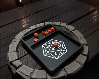 May include: A black dice tray with a white D20 design and several orange dice. The tray is on a dark wooden table, surrounded by a stone border. The scene is dimly lit, suggesting a game night setting.