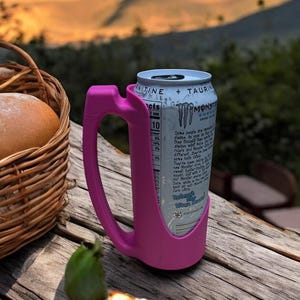 May include: A pink can holder with a Monster Energy drink can inside. The can holder has a handle and is designed to hold a can securely. The can is blue and white with the Monster Energy logo and text. The can holder is sitting on a wooden table with a basket of bread and fruit in the background.