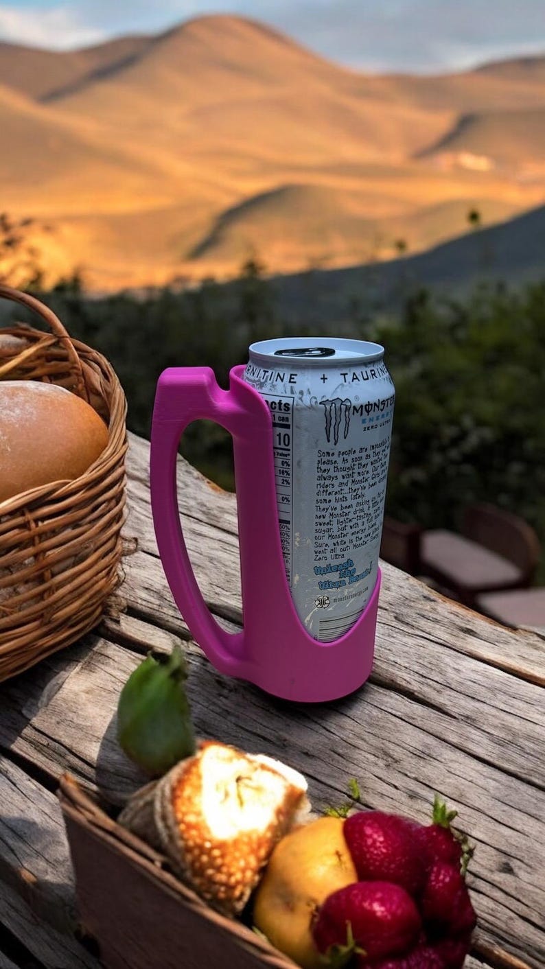 May include: A pink can holder with a Monster Energy drink can inside. The can holder has a handle and is designed to hold a can securely. The can is blue and white with the Monster Energy logo and text. The can holder is sitting on a wooden table with a basket of bread and fruit in the background.