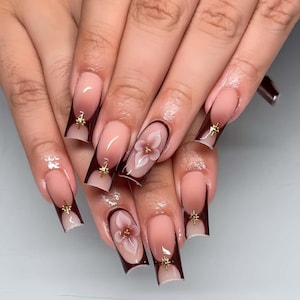 Puede incluir: Uñas artificiales largas y cuadradas con un degradado nude a marrón oscuro. Algunas uñas presentan un diseño floral en 3D y adornos dorados en forma de estrella. Manicura cuidada y acabado brillante.