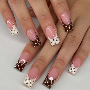 Puede incluir: Una manicura con uñas artificiales largas y cuadradas. Las uñas presentan un diseño de manicura francesa con una base rosa claro y una punta blanca adornada con lunares negros. Algunas uñas tienen una punta marrón oscuro con lunares blancos.