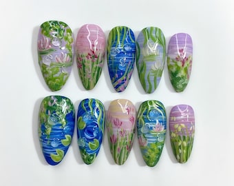 May include: Ten artificial nails with a water lily design. Each nail features a different scene with water, lily pads, and flowers in shades of blue, green, pink, and purple. The nails are oval-shaped and arranged on a white surface.