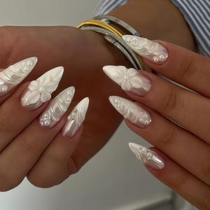 Puede incluir: Uñas acrílicas blancas nacaradas con forma de estilete. Las uñas presentan diseños florales y de remolinos en 3D, acentuados con pequeñas perlas redondas. Una manicura glamurosa y elegante.