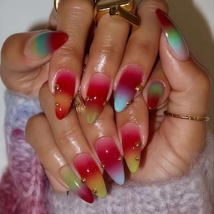 May include: Close-up of a hand with long, stiletto-shaped nails. The nails feature a vibrant gradient design, transitioning from red to yellow, green, and blue. Small gold studs adorn each nail. Several gold rings are also visible.