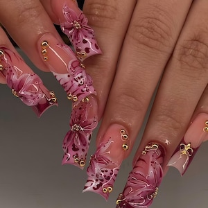May include: Long, sculpted fingernails with a clear base and intricate designs. The nails feature a gradient of pink and burgundy, adorned with 3D floral accents, gold beads, and heart patterns. The overall effect is elegant and artistic.