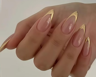 May include: Close-up of long, almond-shaped nails with a nude base and a gold metallic French tip. The nails are well-manicured and reflect light, showcasing a polished and elegant aesthetic. The overall look is sophisticated and stylish.