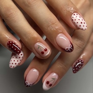 May include: Close-up of a hand with a manicure featuring cherry and polka dot designs. The nails are painted in shades of red, white, and nude, with a glossy finish. Some nails have a cherry design, while others feature white polka dots on a red background.