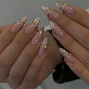 Puede incluir: Primer plano de uñas manicuradas con base nude y detalles dorados. Las uñas en forma de almendra tienen una punta plateada brillante y un diseño dorado en los bordes. Las uñas son largas y elegantes, mostrando un estilo sofisticado.