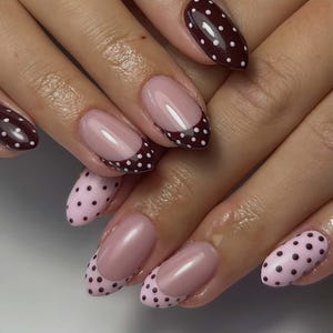 Puede incluir: Primer plano de uñas manicuradas. Algunas uñas son de un borgoña profundo con lunares blancos, mientras que otras son de un rosa suave con un diseño de lunares en las puntas. Las uñas tienen un acabado brillante.
