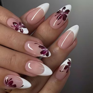 Puede incluir: Primer plano de una mano con uñas largas en forma de almendra. Cada uña presenta una punta blanca, una base transparente y un diseño floral burdeos. Las uñas están manicuradas profesionalmente y muestran un arte de uñas elegante y artístico.