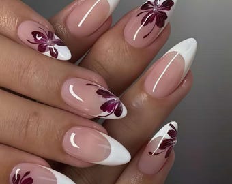 Peut inclure: Gros plan sur une main avec de longs ongles en amande. Chaque ongle présente un bout blanc, une base transparente et un motif floral bordeaux. Les ongles sont manucurés professionnellement et présentent un nail art élégant et artistique.