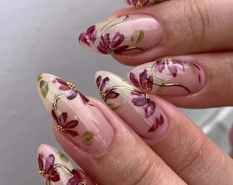Peut inclure: Ongles en amande avec un motif floral. Les ongles sont peints avec une base rose clair et présentent des fleurs bordeaux avec des accents dorés. Des feuilles vertes et de petites perles dorées complètent le design, créant un look délicat et artistique.