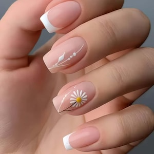 Puede incluir: Primer plano de una mano con uñas manicuradas. Las uñas presentan una base rosa claro con puntas blancas y diseños decorativos. Una uña tiene una margarita blanca con centro amarillo, otras tienen patrones lineales blancos.