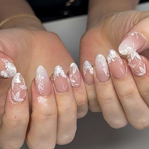 Könnte beinhalten: Mandelförmige Nägel mit nude-farbener Basis und weißen Blumenmustern. Die Nägel zeigen filigrane weiße Blumen- und Blattdesigns mit einem Farbverlauf zu den Spitzen. Das Design ist elegant und für besondere Anlässe geeignet.