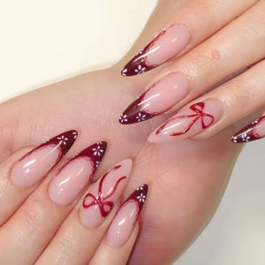 Puede incluir: Uñas almendradas con base nude y puntas burdeos. Las puntas están decoradas con detalles de flores blancas y un diseño de lazo rojo. Las uñas están pintadas con un acabado brillante.