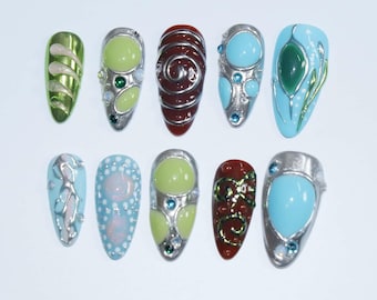 May include: A collection of ten artificial nails with various designs. The nails feature a range of colors, including green, brown, and blue, with silver metallic accents. Some nails have intricate patterns, while others have smooth, solid colors.