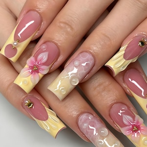 May include: Long square-shaped artificial nails featuring a pink and pale yellow French manicure design. The nails are decorated with three-dimensional pink floral art, small white pearls, gold metallic accents, and clear raised water droplet textures.