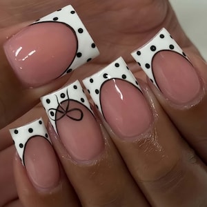 May include: A set of artificial nails with a French manicure design. The nails feature a light pink base with white tips adorned with black polka dots. One nail has a black bow design.