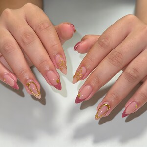 May include: Almond-shaped nails featuring a pink and clear French manicure base with deep pink tips. The design is accented with gold metallic floral decals and small gold studs placed near the cuticles and along the nail beds for detail.