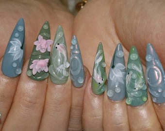 May include: A set of long, pointed acrylic nails with a water-themed design. The nails feature a gradient of blue and green hues, with white floral and koi fish accents, and clear droplet embellishments.