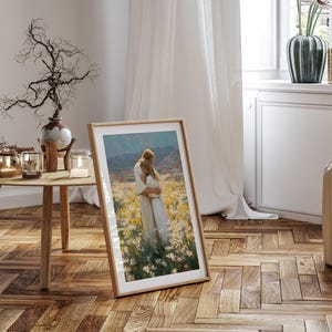 May include: A framed art print featuring a painting of two figures embracing in a field of flowers. The artwork is displayed in a light wood frame, leaning against a wall in a room with a wooden floor and a round wooden table.
