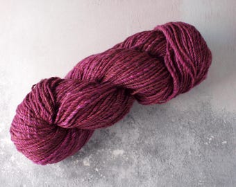 May include: A skein of hand-dyed yarn in shades of deep magenta and burgundy. The yarn is tightly wound, showing the texture and color variations. Ideal for knitting or crochet projects.
