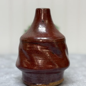May include: A small, hand-thrown ceramic vase with a deep red glaze. The vase has a rounded body and a narrow neck.