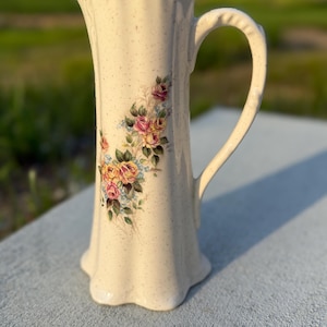 May include: Cream-colored ceramic pitcher with a floral design featuring pink and yellow roses and blue accents. The pitcher has a tall, slender shape with a scalloped rim and base, and a curved handle. The surface has a speckled texture.