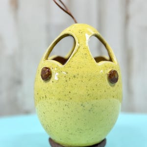 May include: A yellow ceramic egg-shaped hanging planter with two eye-shaped openings and two brown decorative accents. The planter has a brown base and a brown leather hanging loop. The ceramic has a speckled texture.