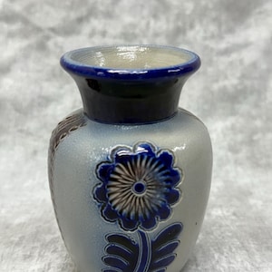 May include: A small, ceramic vase with a blue and white floral design. The vase has a rounded body and a flared rim, with a dark blue band at the top. The flower is a detailed blue and brown design. The vase is approximately 5 inches tall.