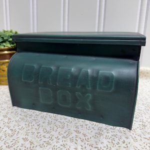 May include: A dark teal bread box with the embossed text "BREAD BOX" on the front. The box has a curved top and a hinged lid. The background includes a white wall and a patterned surface.