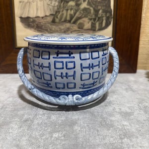 May include: A blue and white ceramic container with a lid. The container has two curved handles and a decorative pattern of squares and symbols. The lid and handles are light blue, and the body is white with blue accents. The container is likely for decorative or storage purposes.