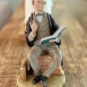 May include: A ceramic figurine of an elderly man seated on a brick structure, holding an open book. The figure wears a suit, bow tie, and a black overcoat. The book has green pages. The figurine is on a beige base.