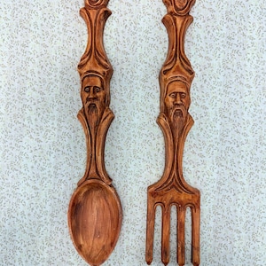 May include: A wooden spoon and fork set with carved faces and ornate handles. The spoon and fork are brown and appear to be made of wood. The set is designed for wall decor.