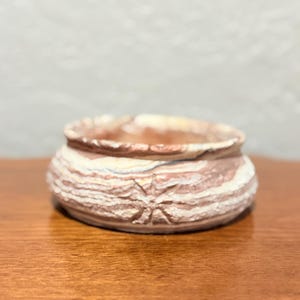 May include: A small, round, ceramic pot with a textured, layered design in shades of brown, cream, and tan. A star-shaped pattern is carved into the side. The pot sits on a wooden surface.