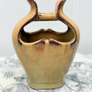 May include: A handcrafted ceramic vase with a unique design. The vase features a tan glaze with brown accents and two curved handles. The open design and color palette make it a decorative piece for any home.