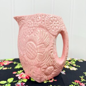 May include: A pink ceramic pitcher with a floral embossed design. The pitcher has a handle and a spout. The floral pattern includes large sunflowers and other stylized flowers. The pitcher is sitting on a black floral tablecloth.