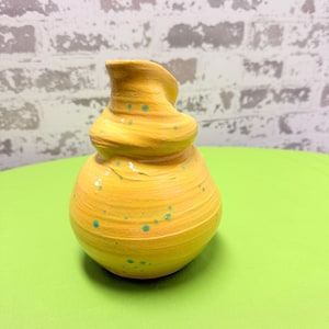 May include: A handcrafted yellow ceramic vase with a unique, layered design. The vase features a bulbous base and a twisted top, with small turquoise-colored speckles. The vase is sitting on a green surface.