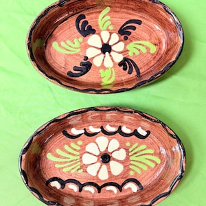 May include: Two oval-shaped, hand-painted ceramic serving dishes. Each dish features a brown base with a floral design in white, black, and green. The rims are dark brown with a scalloped edge. The dishes are displayed on a green background.