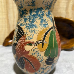 May include: Hand-painted ceramic vase with a beige base and blue speckled accents. The vase features a colorful illustration of a hummingbird and fruit. The bird is red, brown, and black, with a green eye. The fruit is yellow, green, and orange.