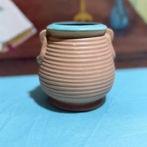May include: A tan ceramic vase with a light blue rim and two small handles. The vase has a ribbed design and sits on a small, round base. The background is blurred, with a blue surface in the foreground.