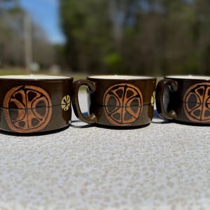 May include: Three brown ceramic mugs with a circular orange and brown design. Each mug has a handle and a cream-colored interior. The mugs are arranged side by side on a light-colored surface.