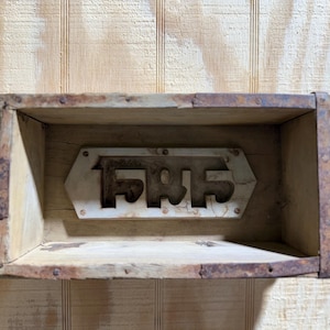 May include: A rustic wooden shelf with a metal accent. The shelf is rectangular with a distressed finish and visible nails. A metal piece with an inscription is mounted on the front. The shelf is mounted on a light wood wall.